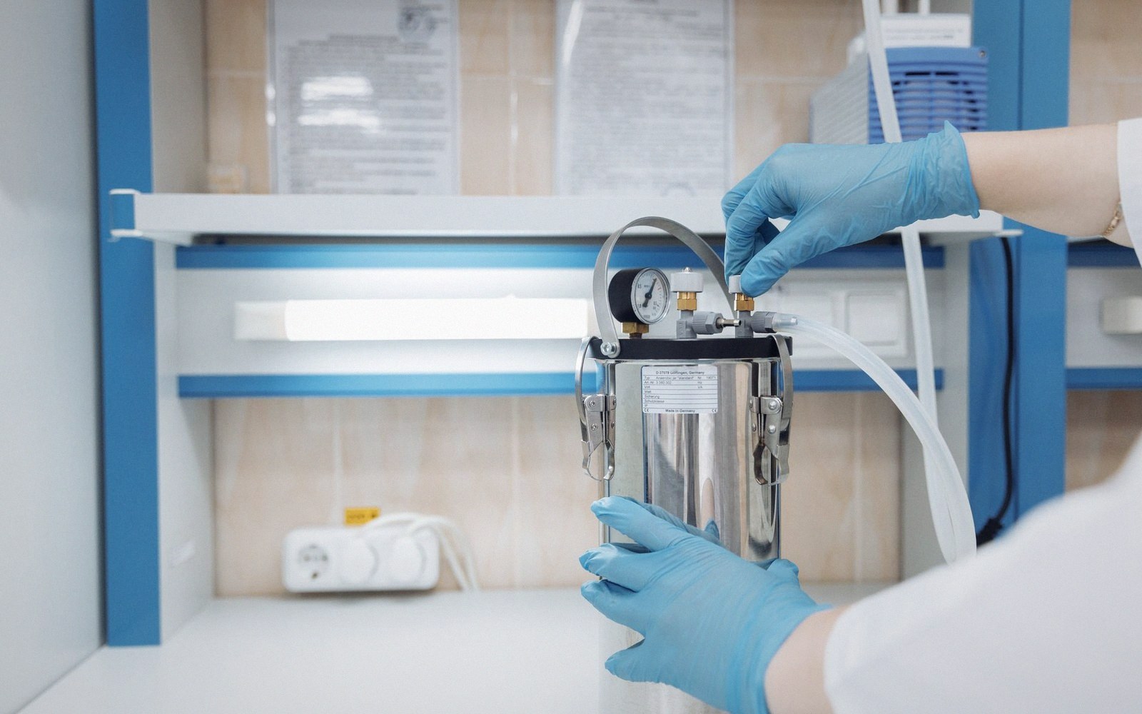 Gloved hands adjust tubing on a pressurized metal canister with a gauge in a lab